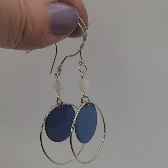 Blue circular drop earrings with silver hoops held by a hand on a white background
