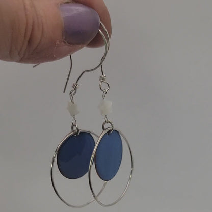 Blue circular drop earrings with silver hoops held by a hand on a white background