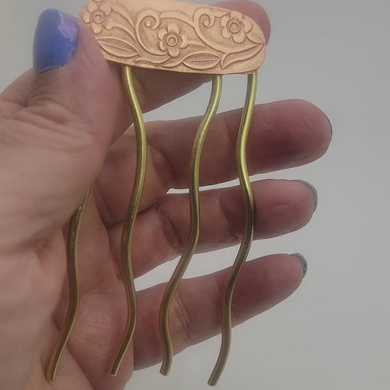 Hand holding a brass and copper hair fork against a white background