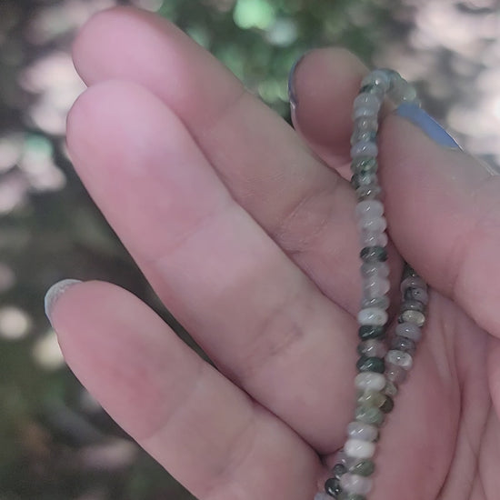 A hand holding a moss agate beaded bracelet in an outdoor environment