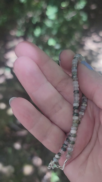 A hand holding a moss agate beaded bracelet in an outdoor environment