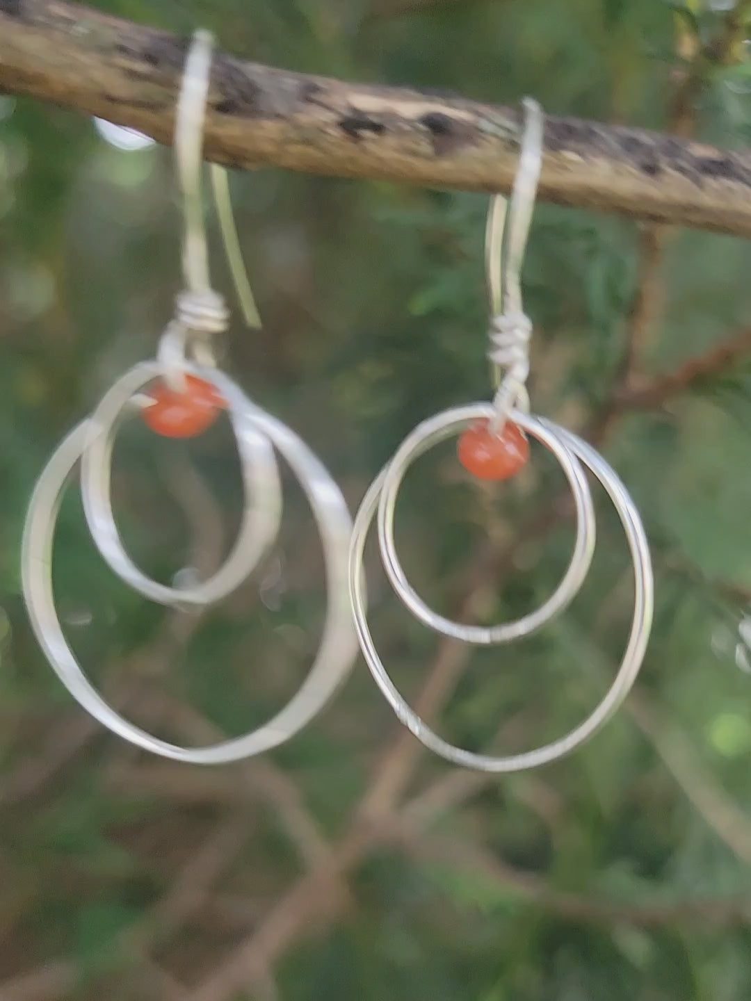 Handcrafted sterling silver hoop earrings with small orange-red carnelian beads in outdoor environment