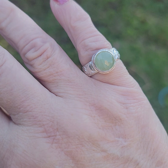 Round Aquamarine gemstone in sterling silver bezel setting with floral patterned band being shown worn on a hand in outdoor environment.