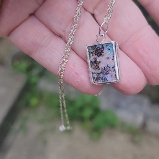A hand holding an iridescent opal pendant with flashes of purple, pink and green in a natural background