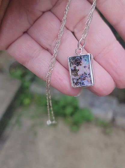 A hand holding an iridescent opal pendant with flashes of purple, pink and green in a natural background