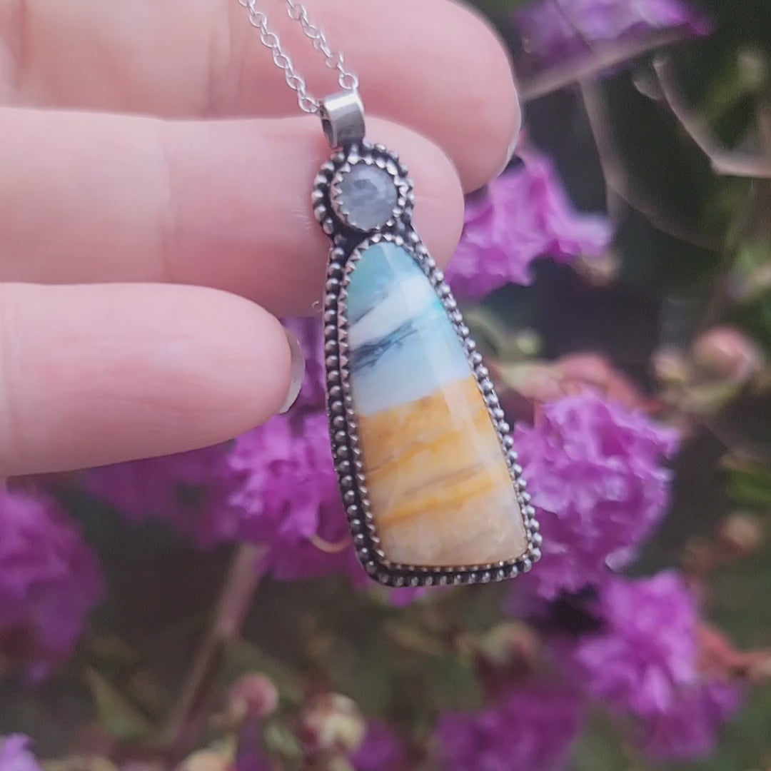 A Scenic opalized wood pendant with aqua blue, cream and golden tan layers in oxidized sterling silver shown in a hand against a natural outdoor environment with a flowering shrub in the background