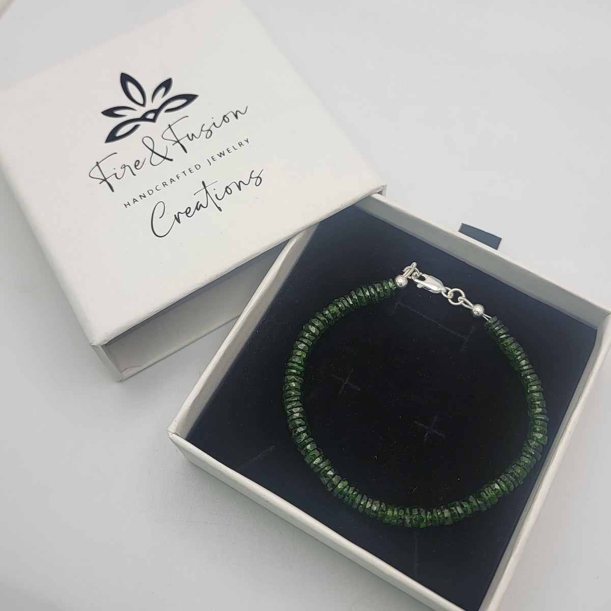 A green beaded bracelet shown in a white branded gift box with a black insert on a white background