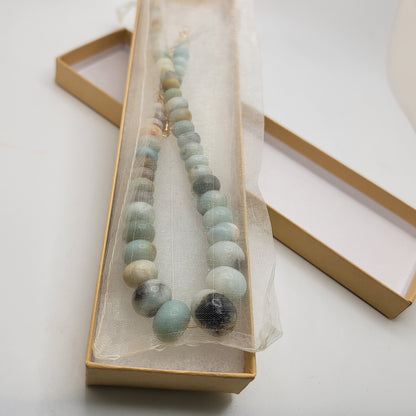 Blue and earthtone beaded gemstone necklace shown in an organza bag in a cardboard gift box on a white background