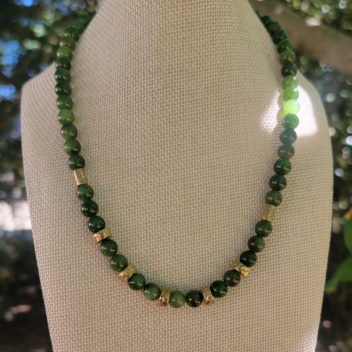 Green beaded necklace with bronze bead accents on a linen bust in an outdoor environment.